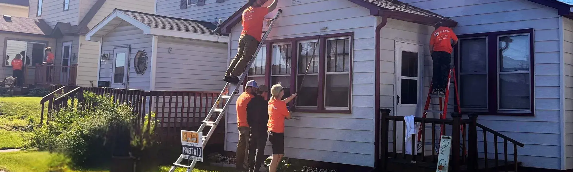 2024 Day of Action volunteers in orange shirts paint the exterior of a Keewatin home.